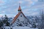 Das Dach der St. Ignatius-Kapelle in Ofterschwang-Schweineberg leuchtet durch Schneewipfel, dahinter Gipfel der Sonnenköpfe östlich des Illertales im Oberallgäu (Januar 2012)